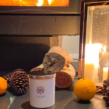 Load image into Gallery viewer, Winter hearth candle in white jar, by fireside with citrus and pine fragrances.
